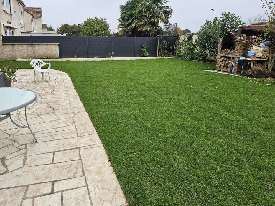 Jardin résidentiel extérieur avec pelouse synthétique verte parfaitement installée, terrasse en pavés de pierre naturelle beige, mobilier de jardin blanc incluant table ronde et chaise